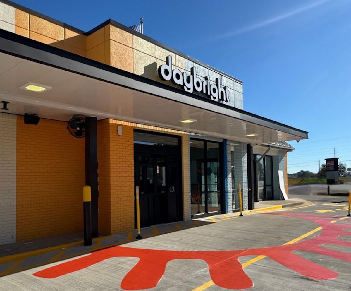 A modern building with a sign reading "daybright" above the entrance, featuring yellow, orange, and white walls, large windows, and a driveway with a sunburst pattern painted on the ground.