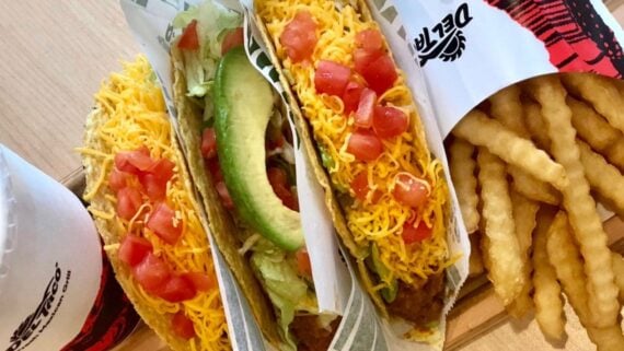 Three hard shell tacos filled with shredded lettuce, ground beef, sliced avocado, diced tomatoes, and shredded cheese, served with crinkle-cut fries and a soft drink in a branded cup.