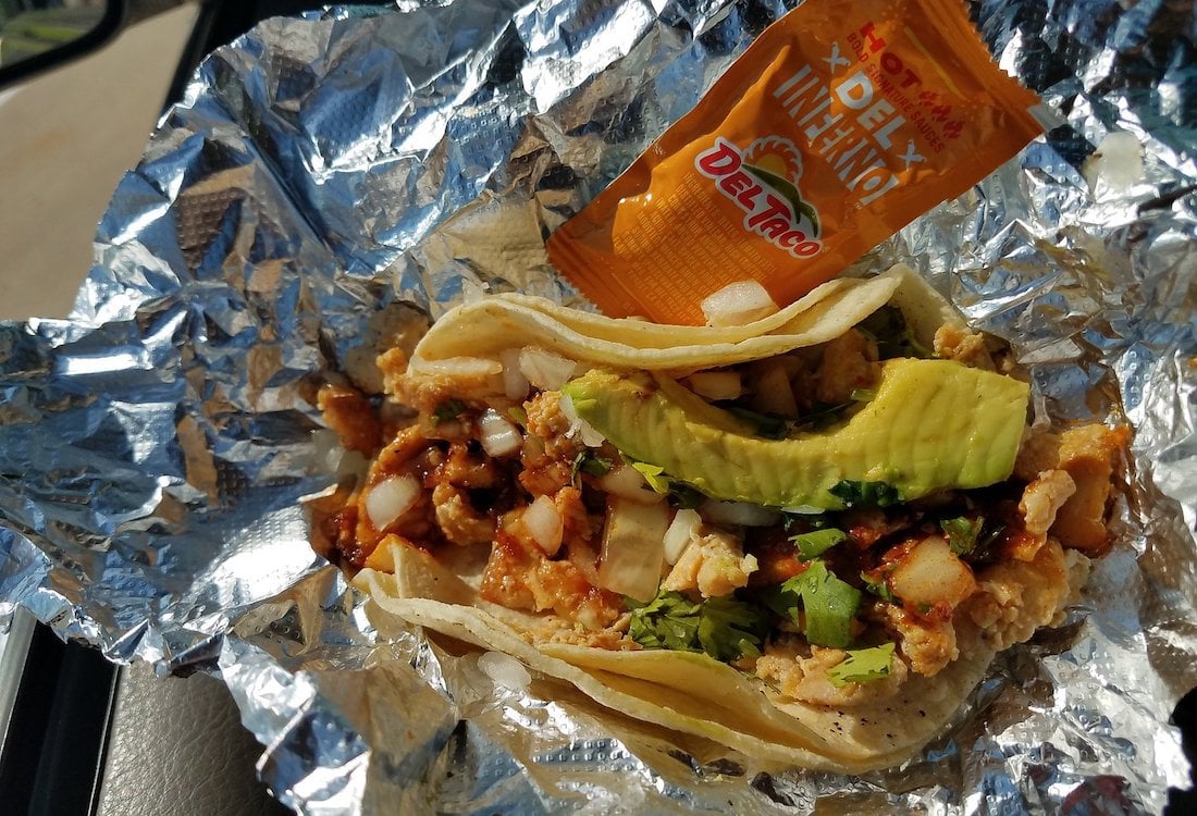 A taco wrapped in foil, filled with scrambled eggs, diced onions, cilantro, and sliced avocado, with a packet of Del Taco hot sauce on top.