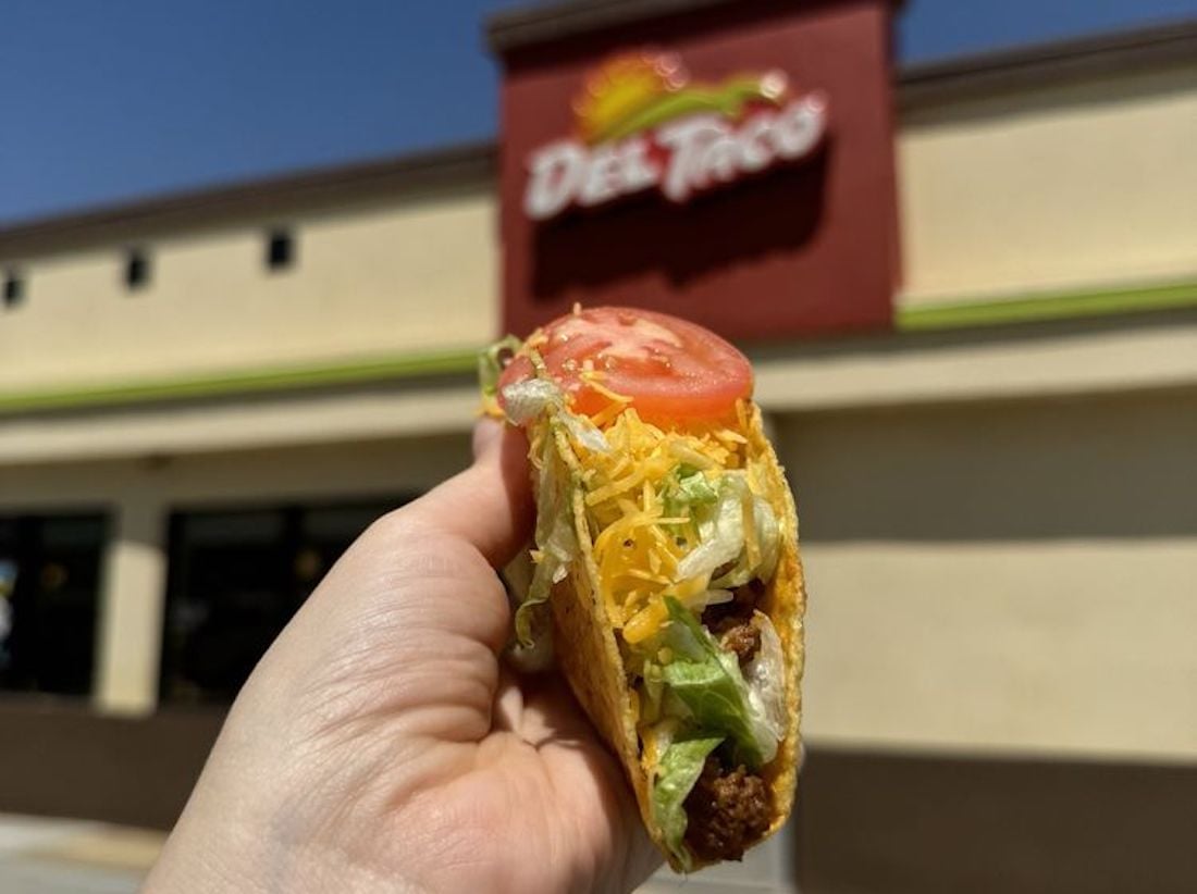 A hand holds up a hard-shell taco filled with lettuce, tomato, cheese, and ground beef in front of a Del Taco restaurant on a sunny day.