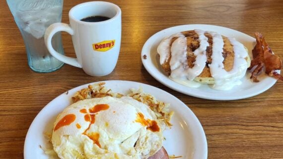 A Denny’s breakfast with hash browns, eggs, toast, pancakes topped with icing, bacon, a mug of coffee, and a glass of ice water, all on a wooden table.