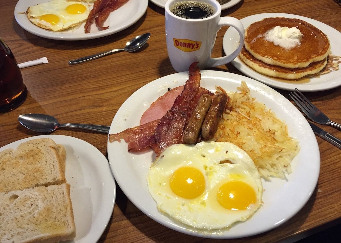 A classic diner breakfast with sunny-side-up eggs, bacon, sausage, ham, hash browns, toast, pancakes with butter, and a cup of coffee on a wooden table at Denny's.