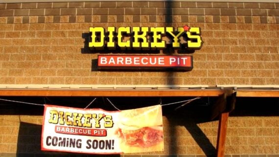 The exterior of a Dickey’s Barbecue Pit restaurant, featuring its bright yellow and red sign on a brick wall, with a banner below that reads “COMING SOON!” and shows an image of barbecue food.