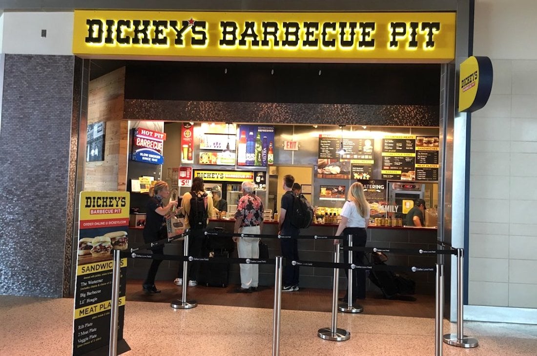 People stand in line at Dickey’s Barbecue Pit, a fast-casual restaurant with a bright yellow sign. The counter displays menus with food options, and there are black queue barriers in front.
