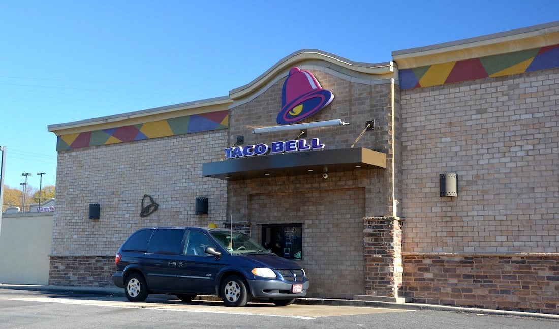 A minivan is parked outside a Taco Bell restaurant with a brick exterior. The Taco Bell logo and sign are above the entrance, and the sky is clear and blue.