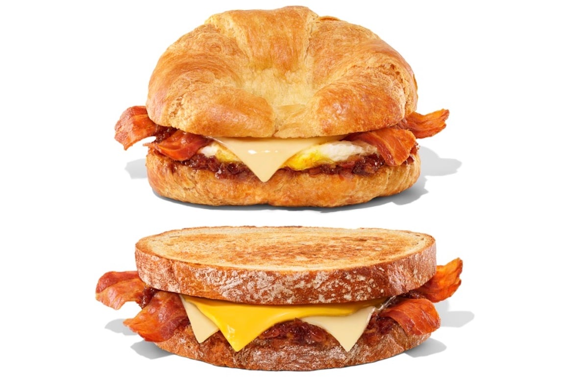 Two breakfast sandwiches: one on a croissant and the other on toasted bread, both filled with bacon, egg, and melted cheese, placed against a white background.