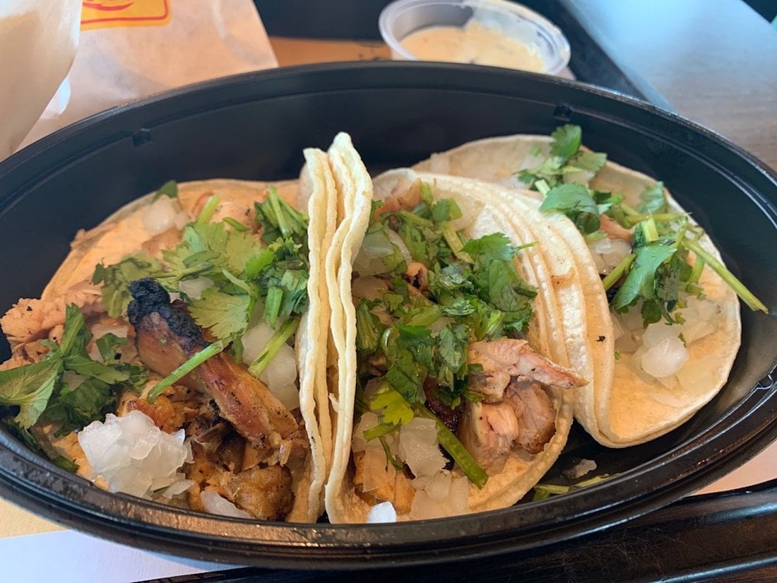 Three street-style tacos filled with grilled meat, chopped onions, and fresh cilantro, served in a black plastic container. A small cup of white sauce is in the background.