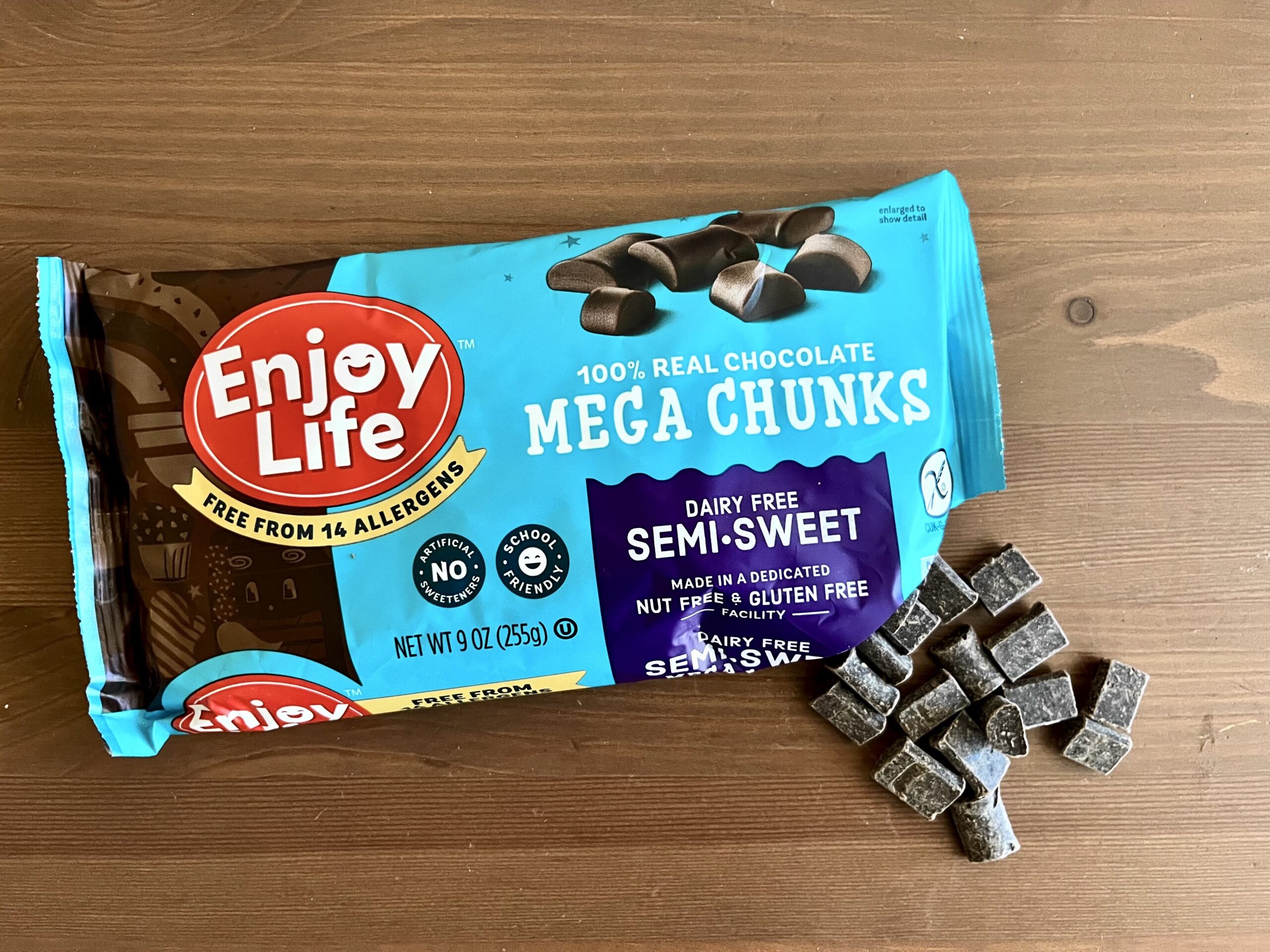 A bag of Enjoy Life Mega Chunks sits on a wooden surface, with several dairy-free semi-sweet chocolate chunks spilled out next to it. The packaging highlights allergen-free and gluten-free features.