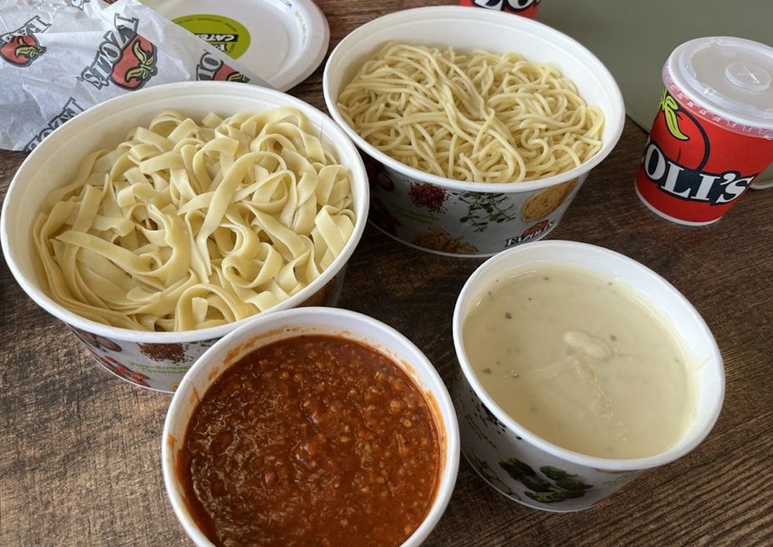 Four containers on a wooden table: one with fettuccine, one with spaghetti, one with creamy white sauce, and one with red meat sauce. Two Oli's branded cups are also visible in the background.