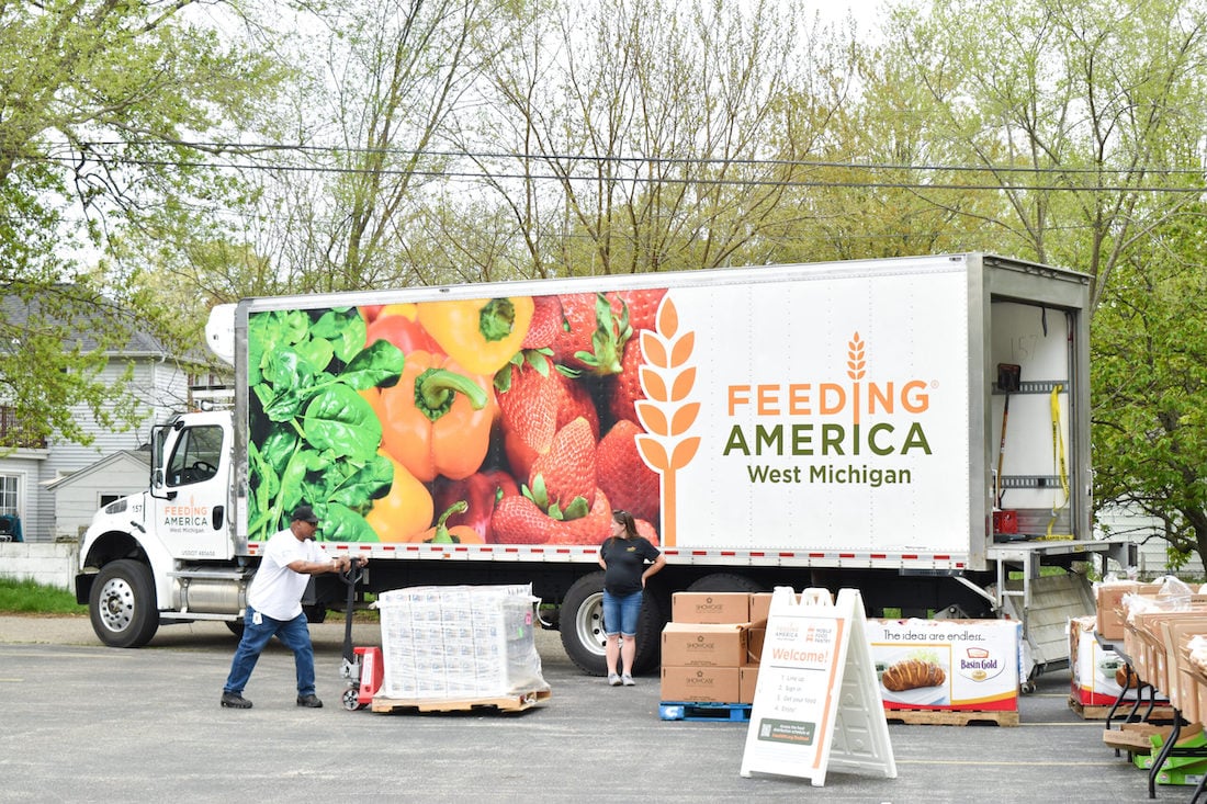 A Feeding America West Michigan truck with images of fresh produce is parked outdoors. Two people unload boxes and supplies near the truck, with tables and signs set up for food distribution. Trees and houses appear in the background.