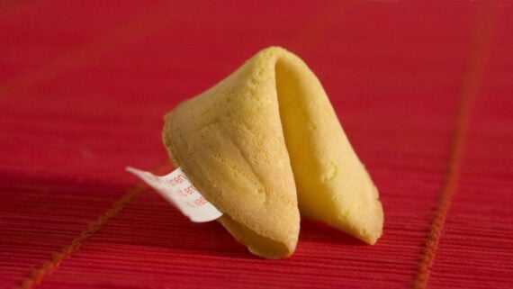 A single fortune cookie with a visible slip of paper peeking out sits on a textured red surface.