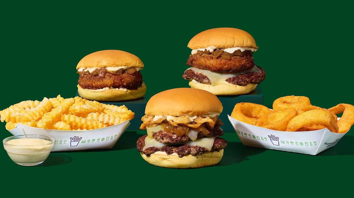 Three cheeseburgers with fried onions and special sauce, from our new fast-food menu, served with crinkle-cut fries, onion rings, and dipping sauce—all arranged on a vibrant green background.