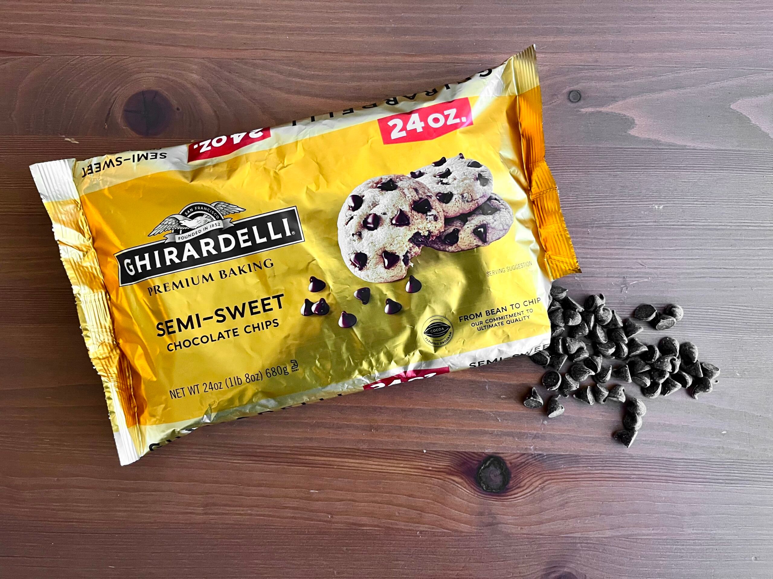 A 24-ounce bag of Ghirardelli semi-sweet chocolate chips lies on a wooden surface with some chocolate chips spilled next to it. The yellow packaging features an image of cookies with chocolate chips.
