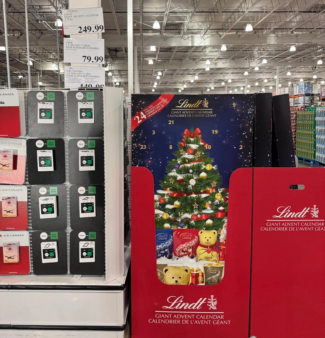 A display in a store features a giant Lindt advent calendar with a decorated Christmas tree and teddy bears next to shelves of gourmet gift cards. Price tags are visible above the display.