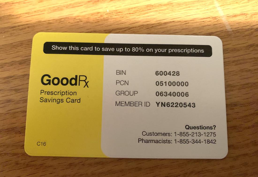 A GoodRx prescription savings card on a wooden surface, showing numbers for BIN, PCN, GROUP, MEMBER ID, and phone numbers for customers and pharmacists. The card is yellow and white.