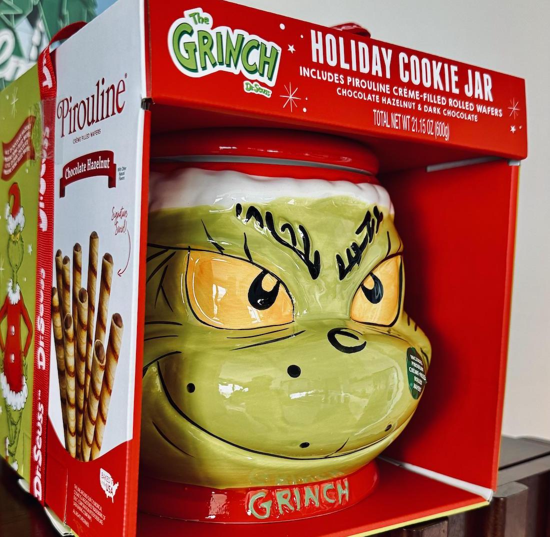 A ceramic holiday cookie jar shaped like the Grinch’s face sits in a festive red box. The set includes Pirouline chocolate hazelnut rolled wafers, shown on the left side of the packaging.