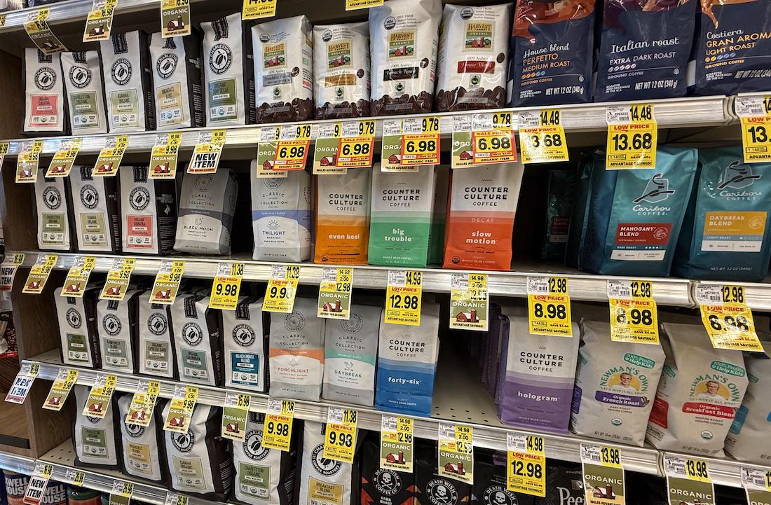 Bags of coffee in various brands and flavors are displayed on grocery store shelves, each with yellow sale price tags. Brands include Counter Culture Coffee and Peet’s Coffee.