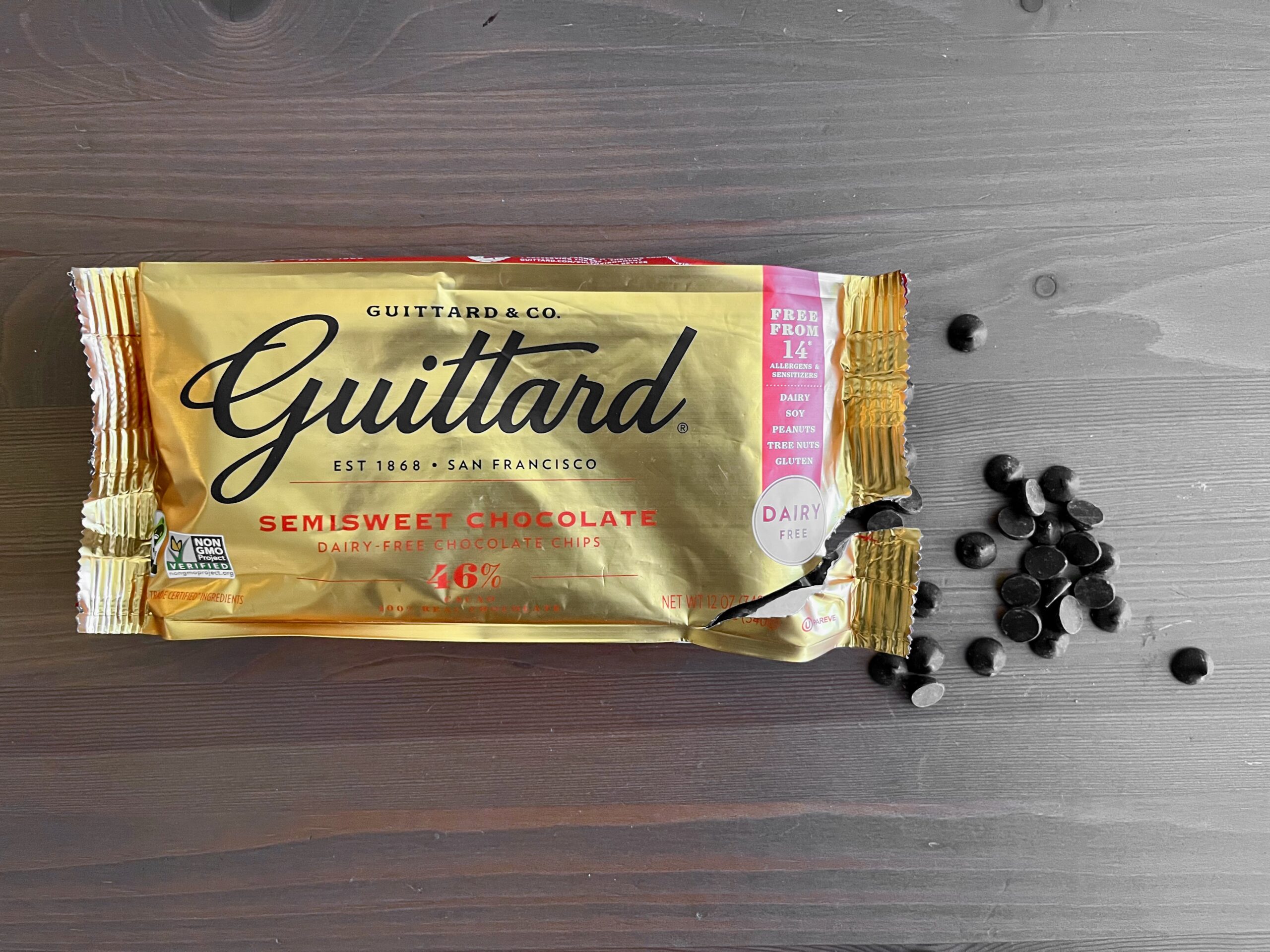 A torn package of Guittard semisweet chocolate chips lies on a dark surface, with several chocolate chips scattered beside it. The gold and red packaging is slightly crumpled.