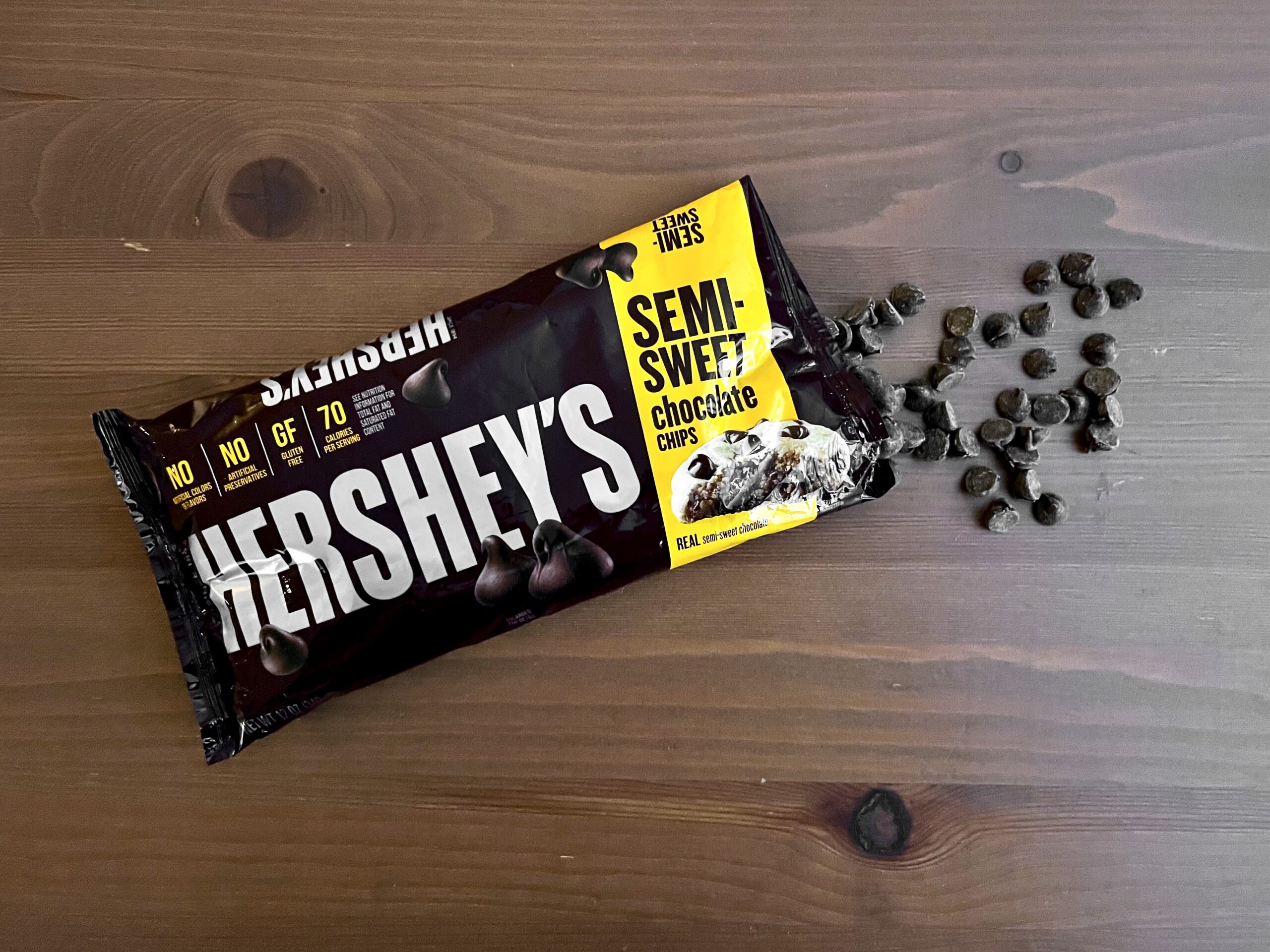 A partially opened bag of Hershey’s semi-sweet chocolate chips lies on a wooden surface, with some chocolate chips spilled out next to the bag.