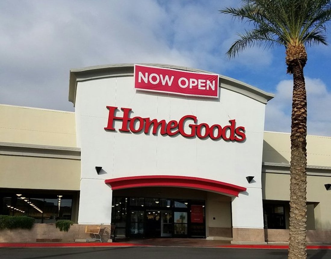 The exterior of a HomeGoods store with a large red "HomeGoods" sign and a red banner above reading "NOW OPEN." A palm tree stands to the right of the store entrance under a partly cloudy sky.