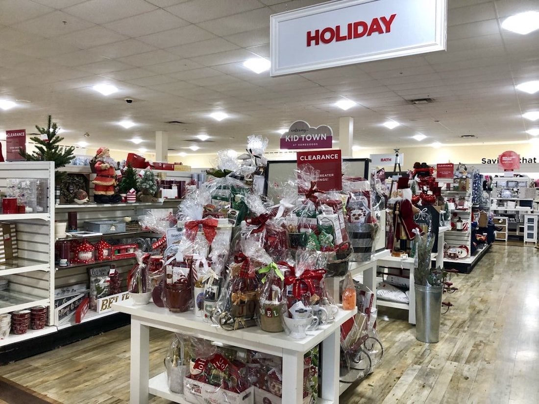 A store display features holiday-themed items, including gift baskets, ornaments, and decorations. A sign reading "HOLIDAY" hangs above, with shelves of festive goods and a clearance section in the background.