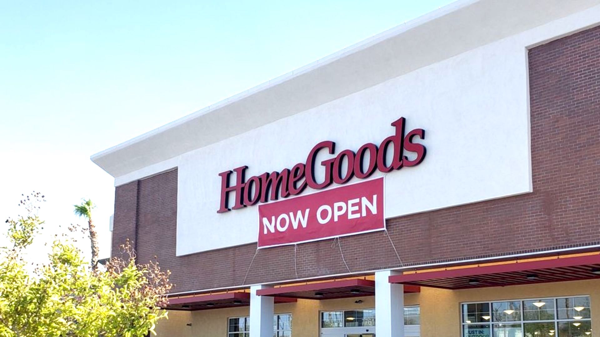 The exterior of a HomeGoods store with a large red and white "NOW OPEN" banner hanging below the store's sign on the building. Trees and part of the sky are visible in the foreground.