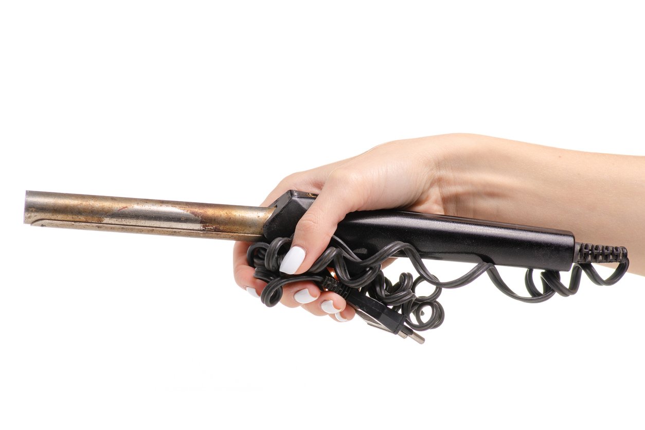 A hand with white-painted nails holds a curling iron with a tangled power cord against a white background.