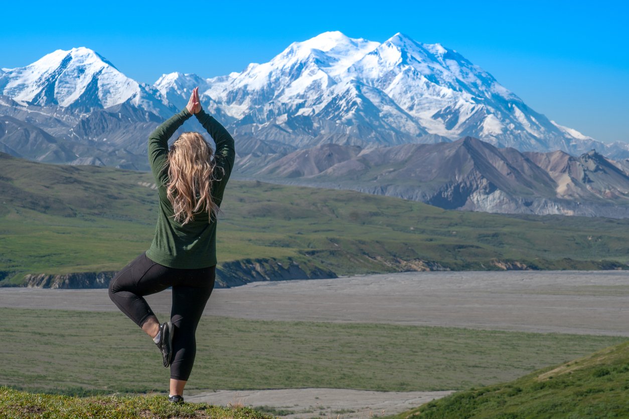 A person with long blonde hair practices a yoga tree pose on a grassy hill, facing snow-capped mountains under a clear blue sky.
