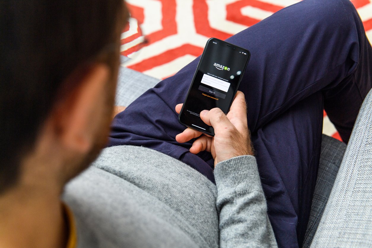 A person sitting on a couch holding a smartphone, logging into an Amazon account. The screen displays the Amazon login page, and the background shows a red and white patterned rug.