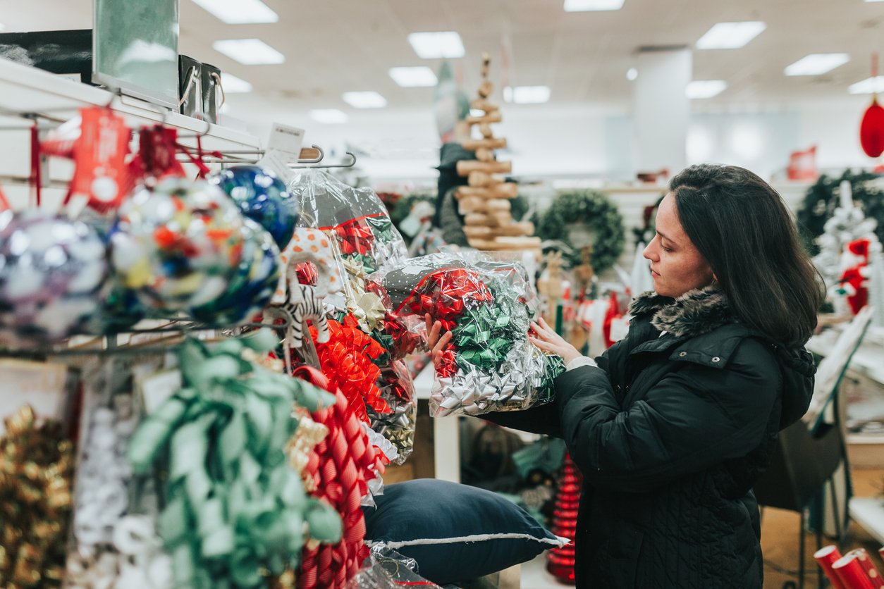 A woman wearing a black coat looks at a package of colorful gift bows in a store aisle decorated with holiday items, including wreaths and a wooden Christmas tree.