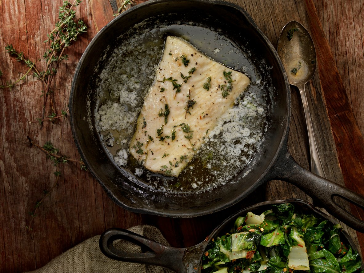 A piece of white fish fillet cooking in a cast iron skillet with melted butter and herbs, on a rustic wooden table, with a side of sautéed greens in a small pan nearby.