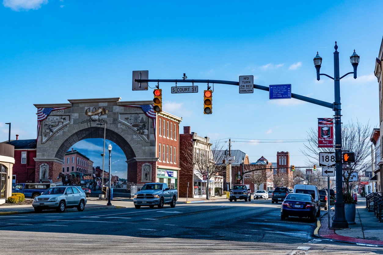A city street scene shows cars stopped at a red light near an ornate arched mural depicting historic buildings, with blue sky above and various street signs and buildings along the road.