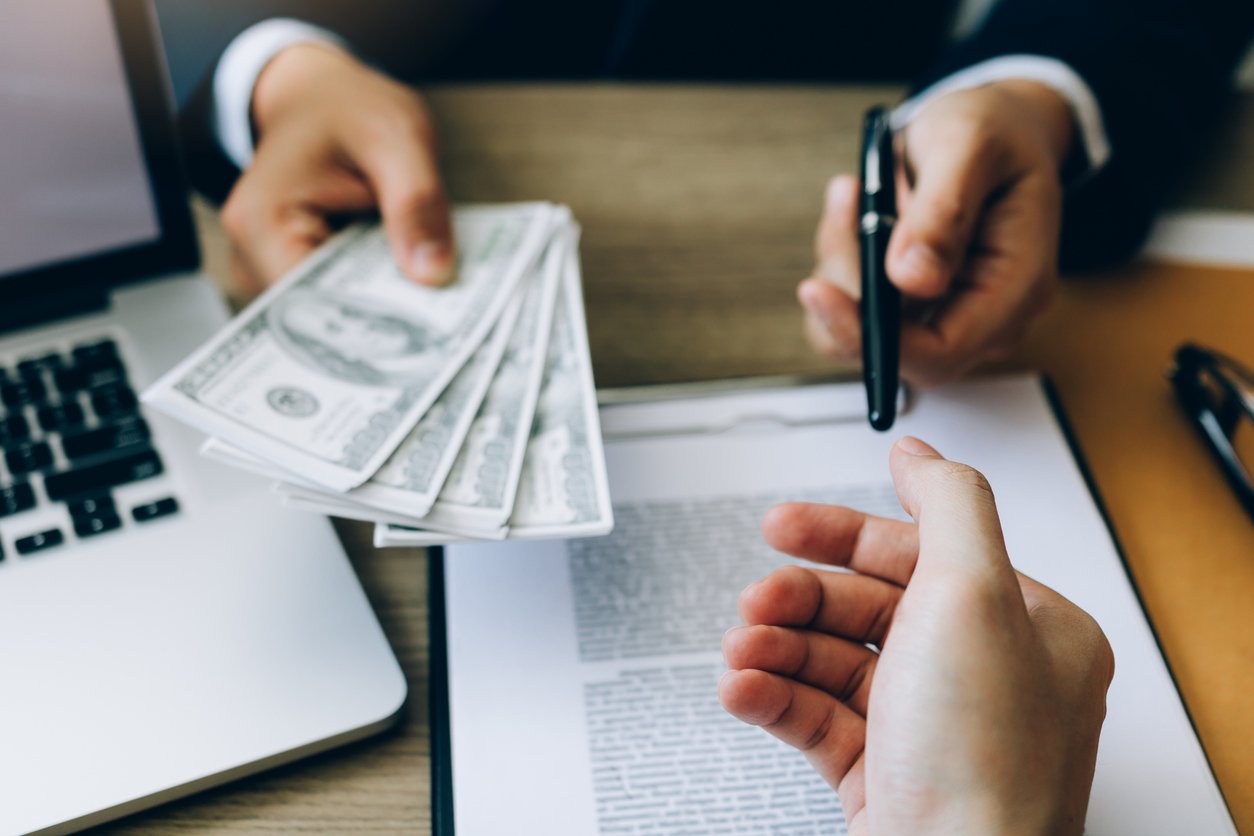A person hands several US $100 bills to another person, who is holding a pen and a document on a clipboard. A laptop and documents are on the desk, suggesting a financial or business transaction.