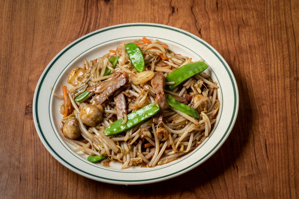 A plate of stir-fried noodles with beef, snow peas, mushrooms, bean sprouts, carrots, and sauce, served on a white dish with a green rim, placed on a wooden table.