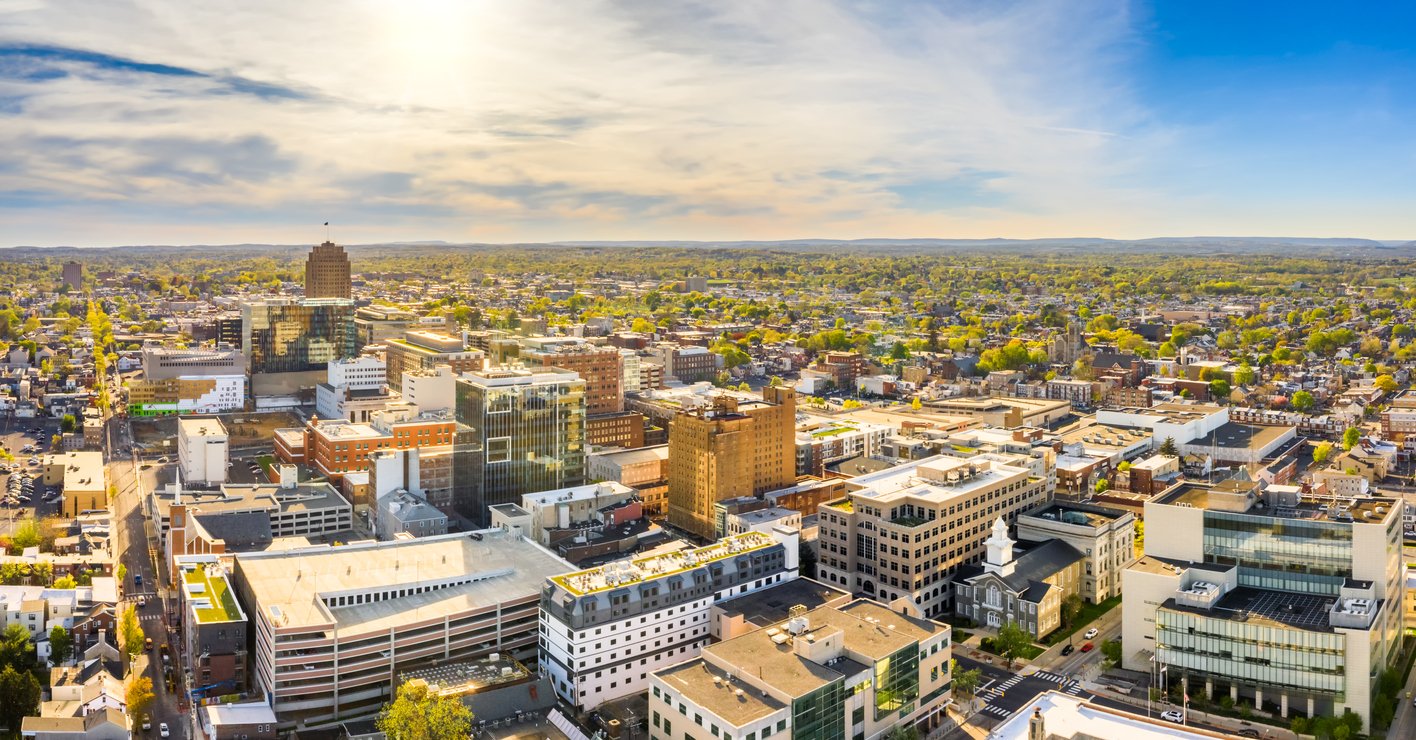 Aerial view of a cityscape on a sunny day, showing a mix of modern and older buildings, streets, and green areas extending into the distance under a blue sky with scattered clouds.