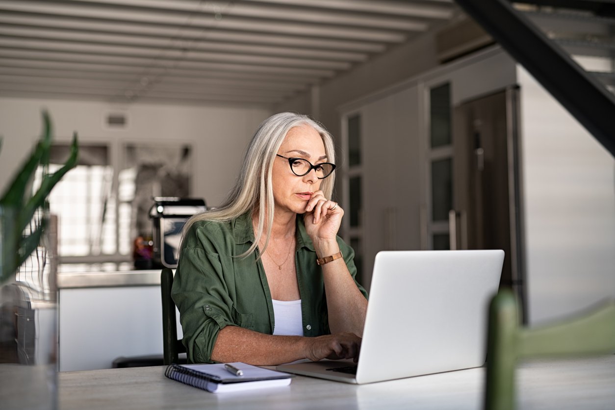 A woman with long gray hair and glasses sits at a table in a modern kitchen, looking thoughtfully at a laptop screen. A notebook and pen are next to her on the table.