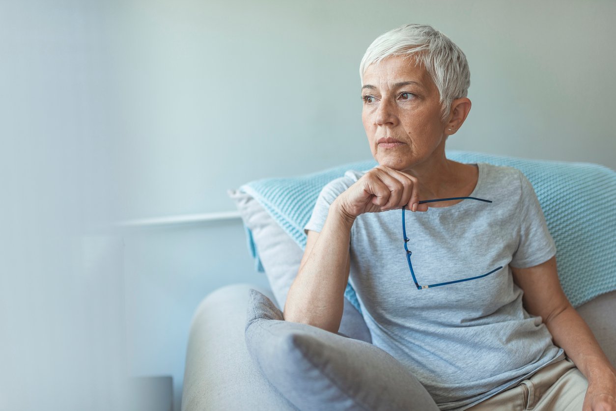 An older woman with short gray hair sits on a couch, holding her glasses and looking thoughtfully into the distance. She wears a light gray shirt and there is a blue pillow behind her.