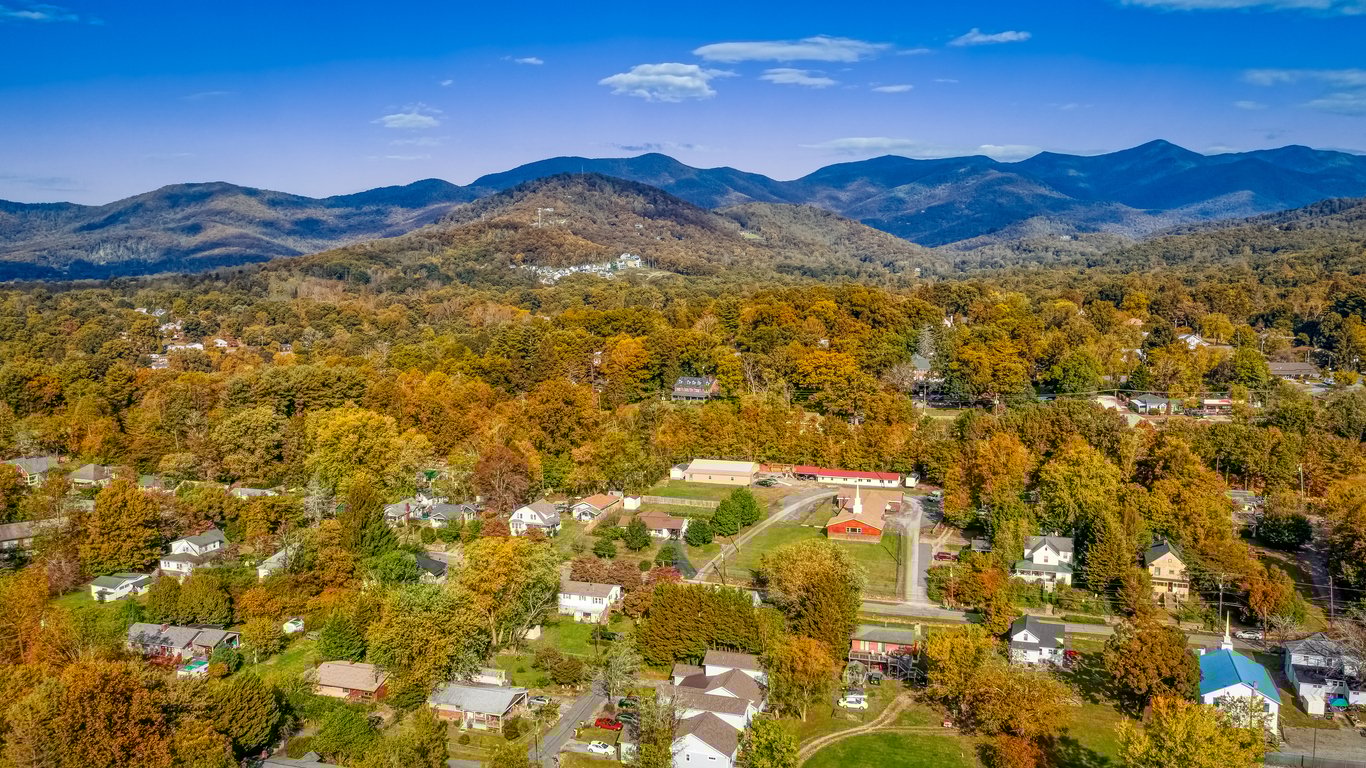 Aerial view of a small town surrounded by trees with autumn foliage, set against a backdrop of rolling mountains under a clear blue sky.