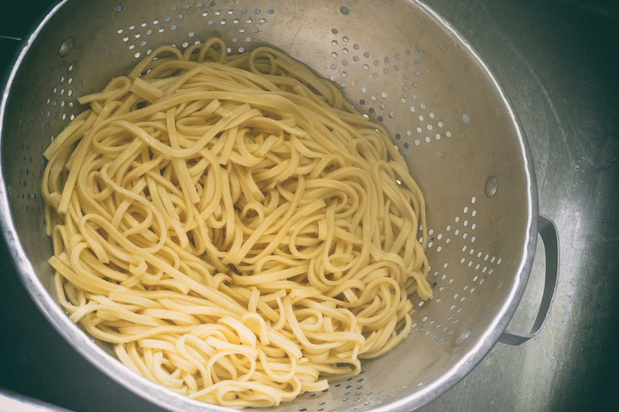 Cooked linguine pasta draining in a metal colander placed in a sink. The pasta appears fresh and yellow, and the colander has small holes for water to escape.