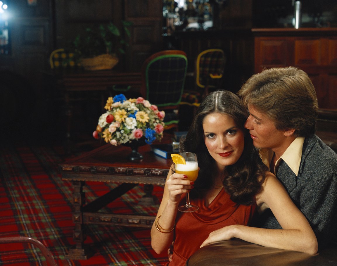 A woman in a red dress holds a cocktail with an orange slice, sitting closely with a man in a suit. They are in a dimly-lit, elegant room with plaid carpet and a floral arrangement on a wooden table behind them.