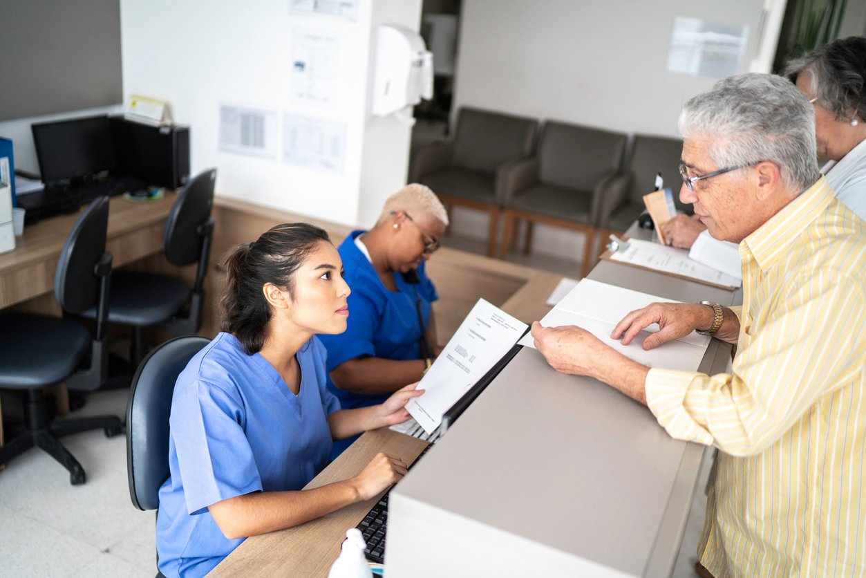 A nurse at a reception desk assists an older man holding paperwork in a medical office, while another staff member works in the background. There are empty chairs and computers nearby.