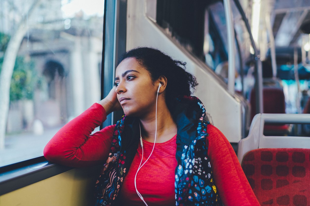 A woman in a red top sits on a bus, resting her head on her hand and looking pensively out the window. She wears earphones and appears deep in thought. The bus seats and window are visible around her.
