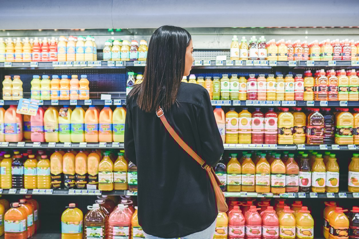 A woman with straight dark hair, wearing a black top and a crossbody bag, stands in front of a supermarket refrigerated section filled with various bottles of juice.