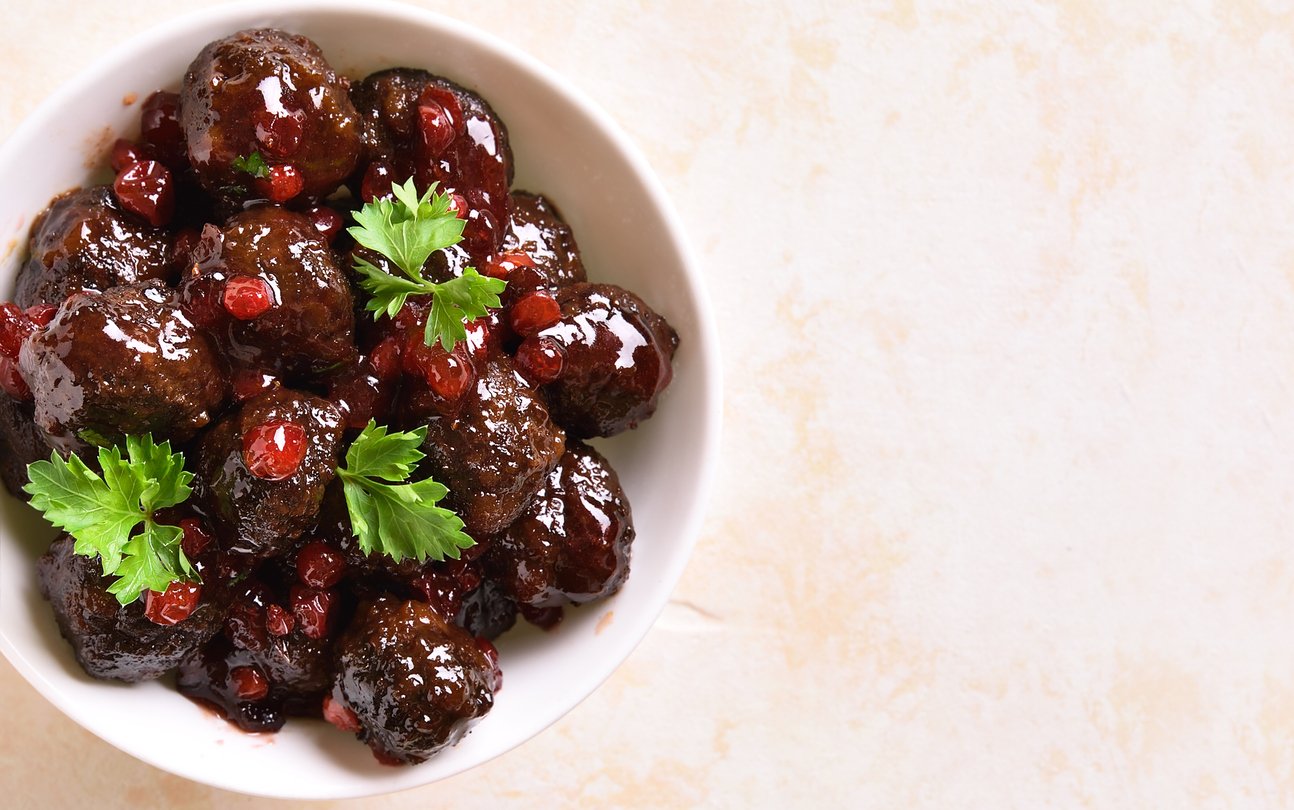 A white bowl filled with glazed meatballs topped with red berries and garnished with fresh parsley, placed on a light-colored surface.