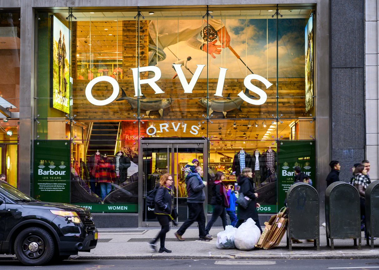 People walk past the glass storefront of Orvis, an outdoor retail store, with large signage and product displays visible inside. A few pedestrians and a parked car are on the street in front.