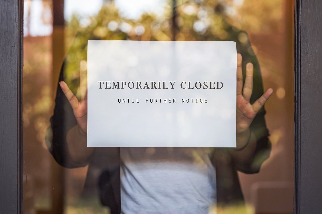 A person holds a sign on a glass door that reads "TEMPORARILY CLOSED UNTIL FURTHER NOTICE," with sunlight and trees reflected in the background.
