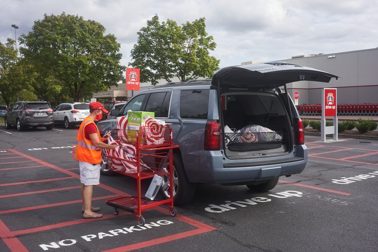 A store employee loads shopping bags into the trunk of a dark SUV parked in a drive-up pickup area outside a retail store. The employee wears an orange safety vest and cap. Red "drive up" signs are visible.