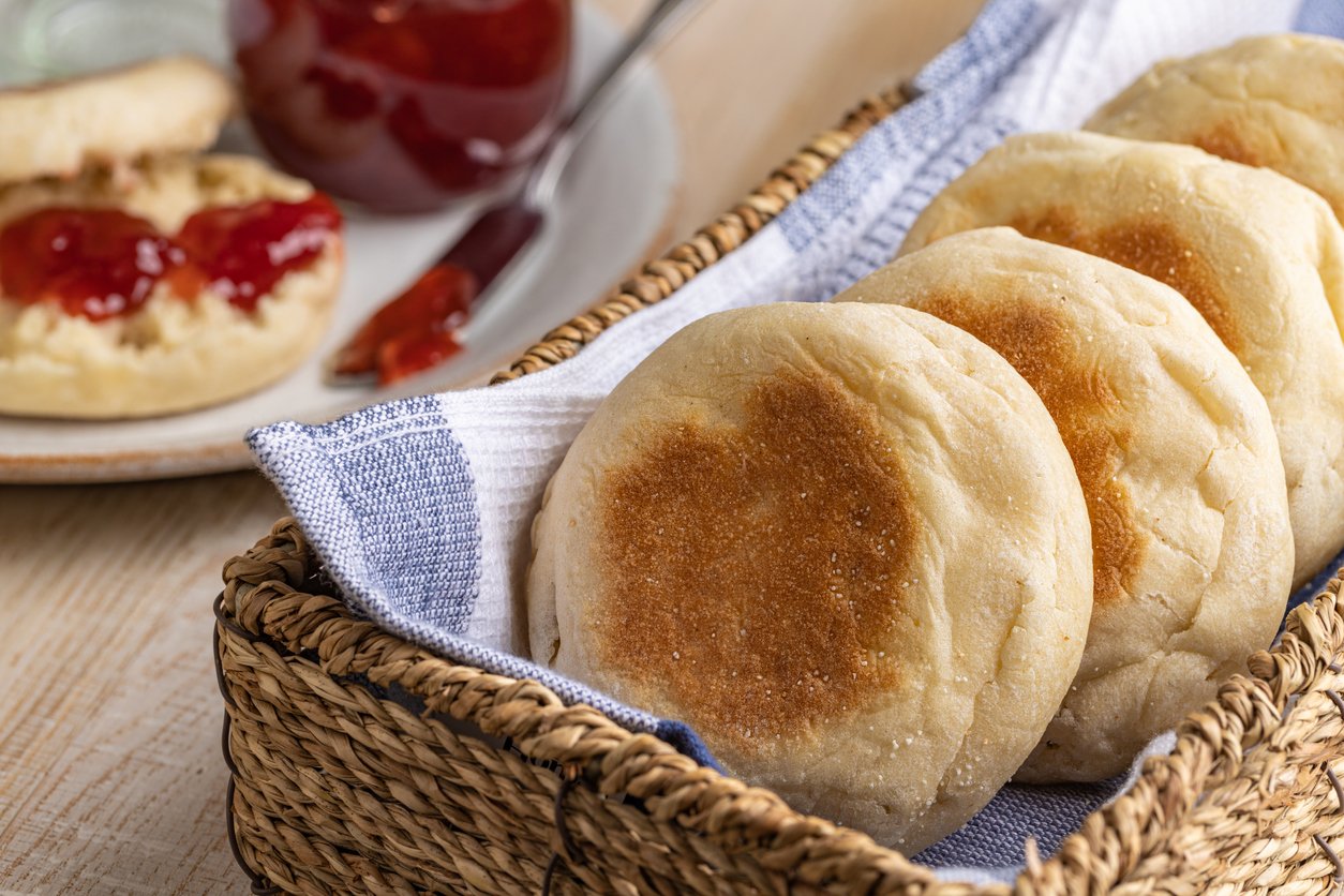 A basket lined with a blue and white cloth holds four English muffins. In the background, a plate has a split muffin topped with red jam, next to a jar of jam with a spoon.