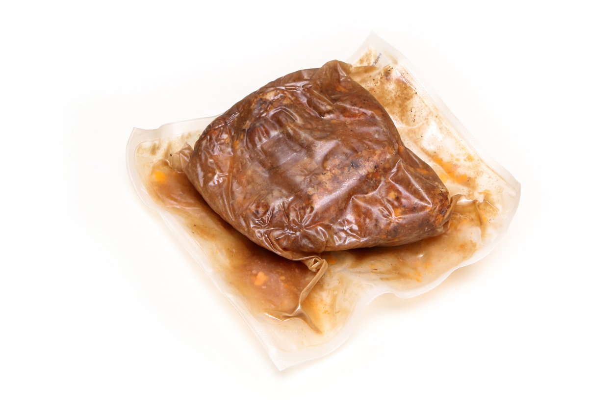 A piece of cooked meat in brown sauce vacuum-sealed in a clear plastic bag, with some sauce pooling inside, on a white background.