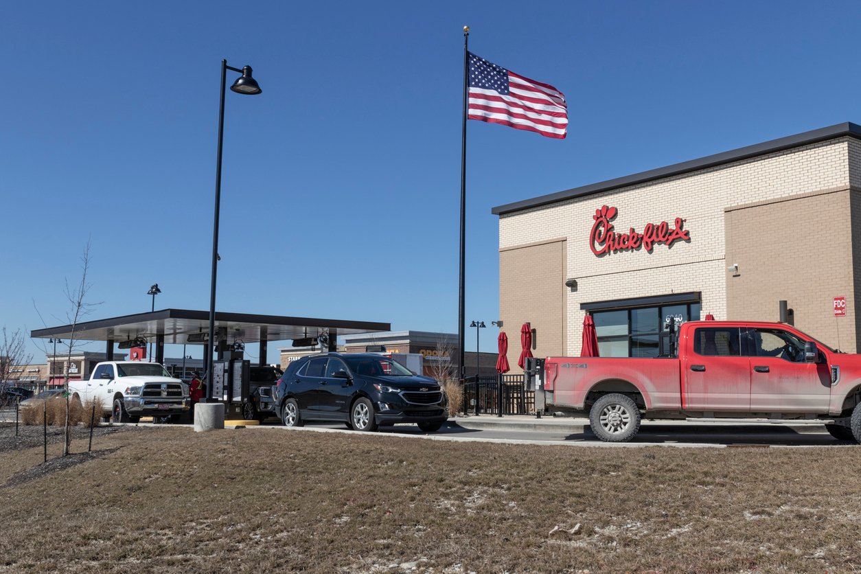 Cars line up at a Chick-fil-A drive-thru on a clear day, with an American flag flying on a pole and a red truck parked in front of the restaurant.